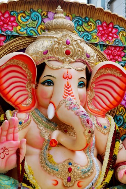 The image depicts a large, colorful statue of the Hindu deity Ganesha, featuring a detailed face with prominent trunk, large ears, and decorative crown. The statue is adorned with ornate jewelry and v