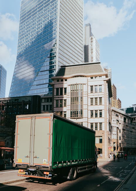 A large green delivery truck with a closed cargo area is parked on a city street in front of tall modern glass skyscrapers and historic stone buildings in the background. The truck is positioned next