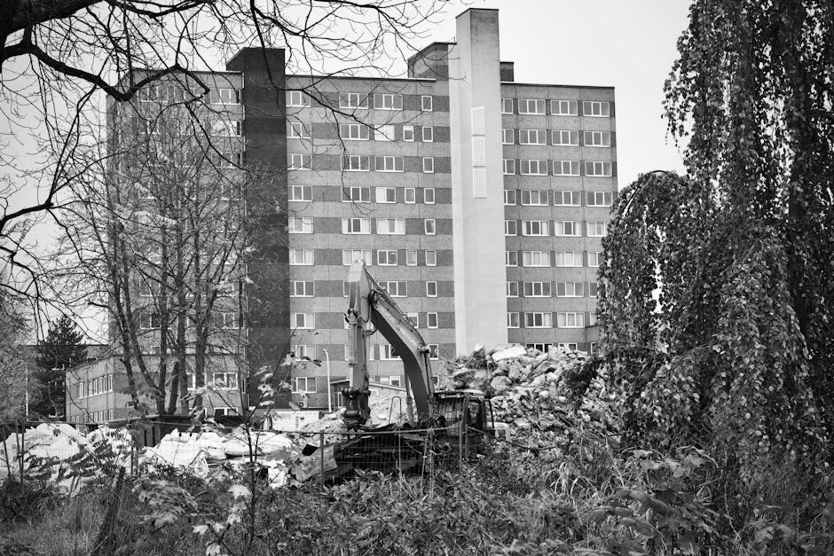 A construction excavator with a long arm and bucket attachment is positioned amidst a pile of rubble and debris on a patch of land adjacent to a multi-storey residential building. The debris consists 
